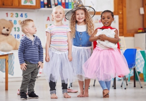 Four children standing in a classroom, each child wearing a costume, they are smiling and laughing. 