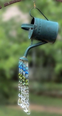 A watering can hanging from a string with strings of beads dangling from it's spout, imitating water.